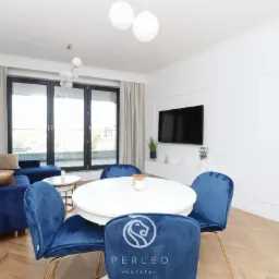 Nowoczesny salon z granatową sofą, okrągłym białym stołem otoczonym welurowymi krzesłami na złotych nóżkach, parkiet w jodełkę i balkon z widokiem na miasto.