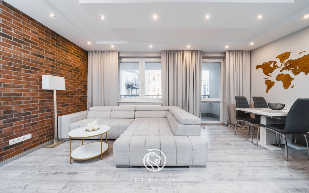 Jasne wnętrze salonu z ceglaną ścianą, narożną sofą, stolikiem kawowym, stołem jadalnym z krzesłami i drewnianą mapą świata na ścianie.