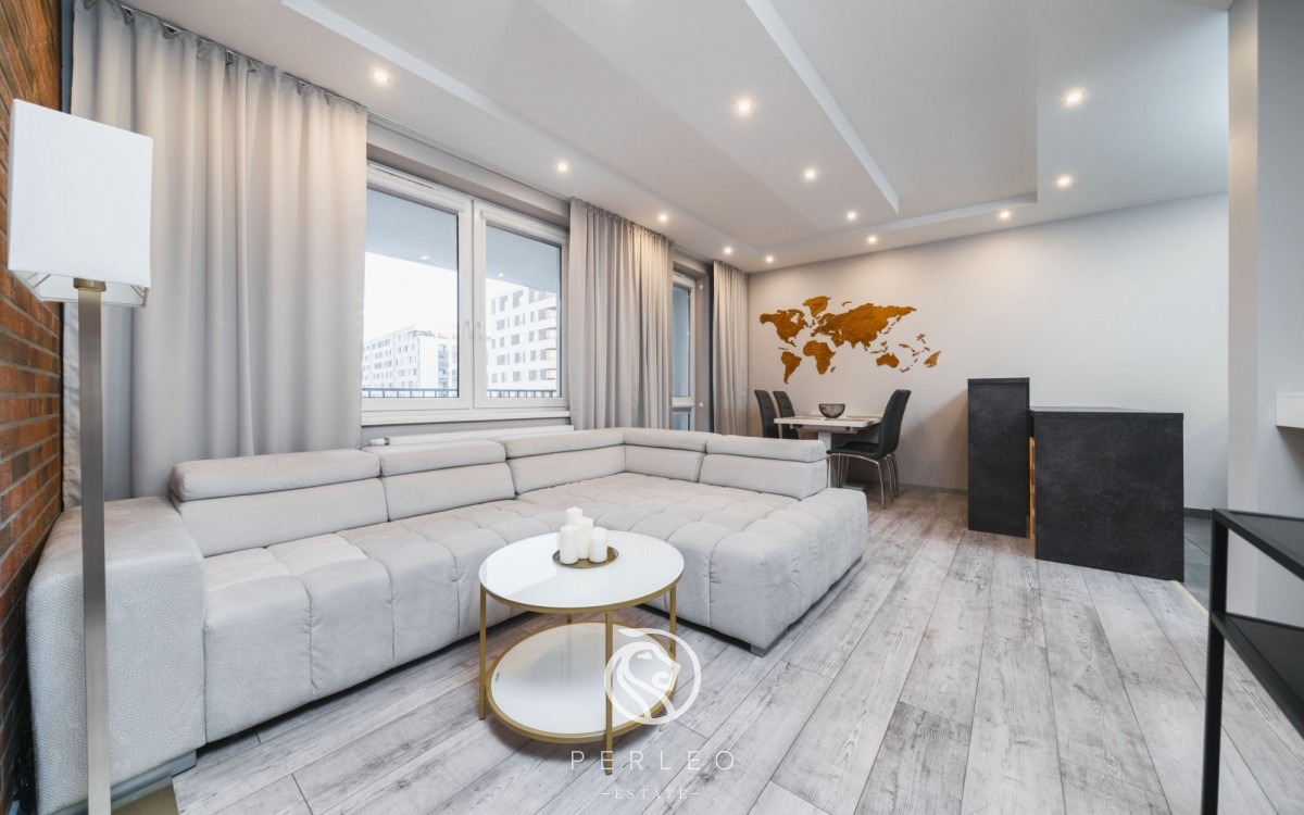 Jasny salon z narożną sofą, okrągłym stolikiem z dekoracjami, drewnianą mapą świata na ścianie i podłogą imitującą bielone drewno.