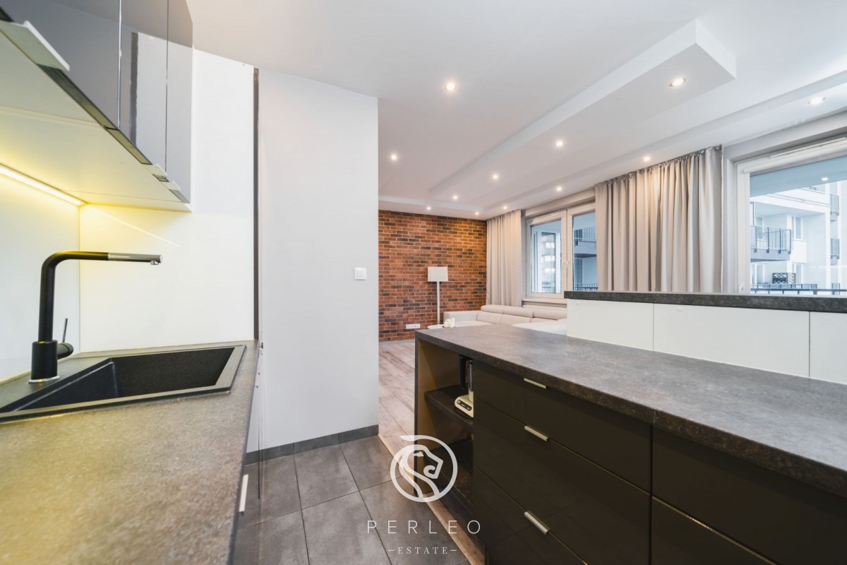 Nowoczesne wnętrze mieszkania z otwartą kuchnią, widoczny czarny zlew i bateria, szare płytki podłogowe, ceglana ściana w tle i logo agencji nieruchomości w dolnej części kadru.