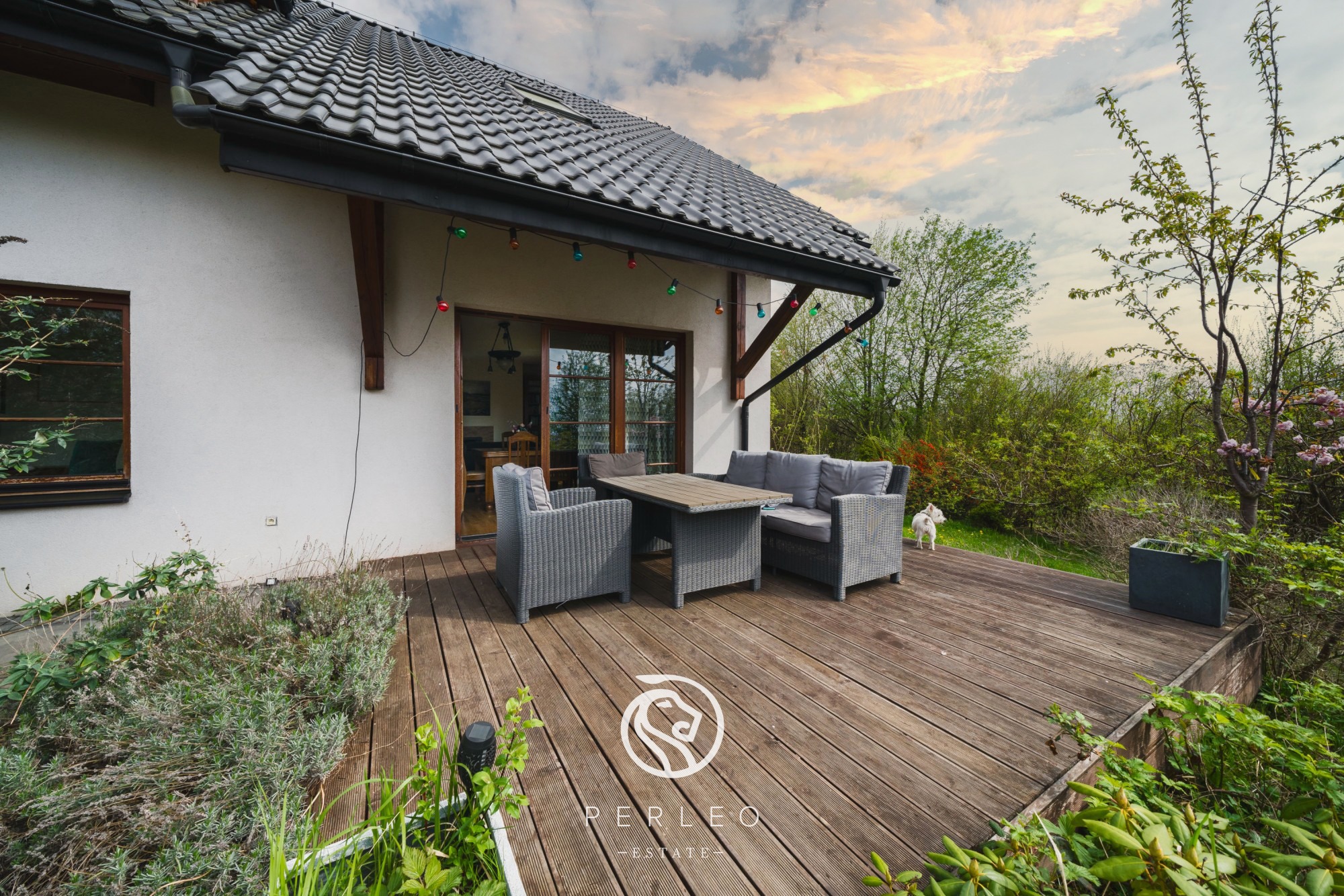 Drewniany taras przy domu z szarymi meblami ogrodowymi, oświetlony girlandą żarówek, z widokiem na ogród i zachodzące słońce.