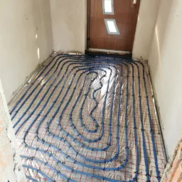 Niebieskie rury ogrzewania podłogowego, ułożone na folii z czerwoną siatką, zamocowane czarnymi spinkami, widoczne ściany w surowym tynku i brązowe drzwi wejściowe