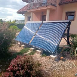 Dwa kolektory słoneczne rurowe zamontowane na zewnątrz domu, otoczone zielenią i kamieniami, z widocznym fragmentem elewacji budynku w tle.