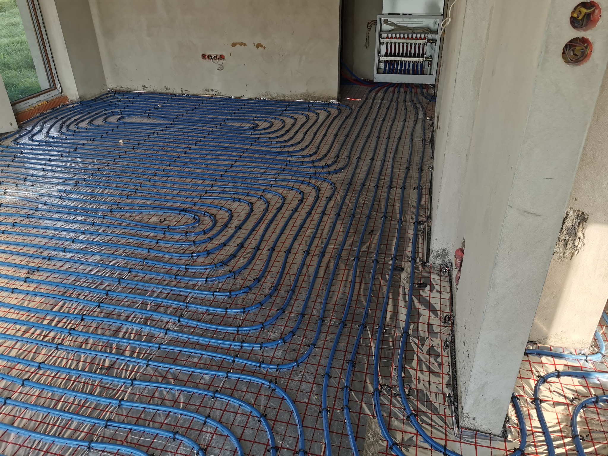 Niebieskie rury ogrzewania podłogowego ułożone na siatce w surowym pomieszczeniu, widoczny rozdzielacz i fragment okna.