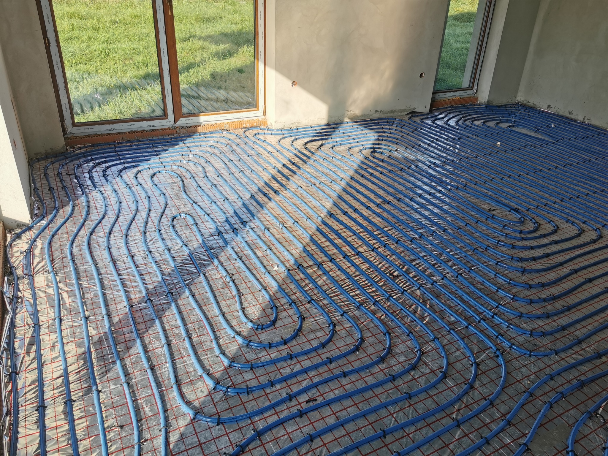 Niebieskie rury ogrzewania podłogowego ułożone na folii z czerwoną siatką w surowym wnętrzu przed wylewką, widok z góry w kierunku okna.