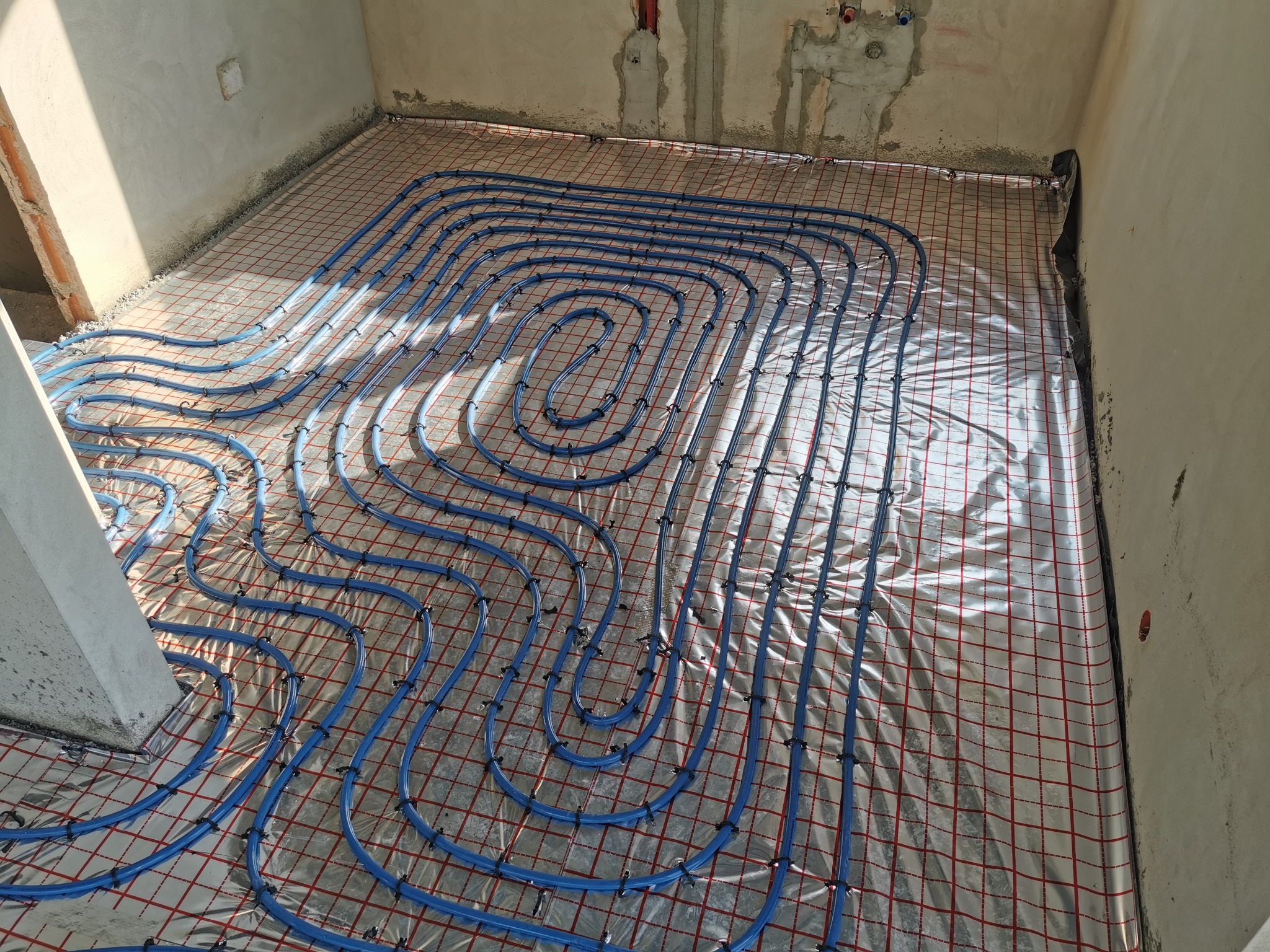 Niebieskie rury ogrzewania podłogowego ułożone na srebrnej folii z czerwoną siatką w surowym wnętrzu, widok z góry na instalację przed wylewką.