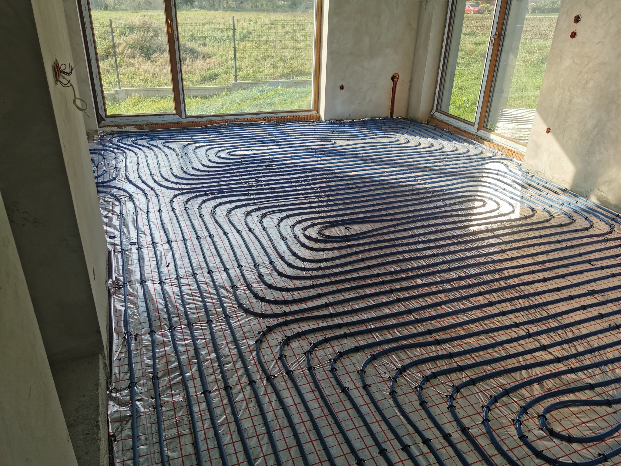 Niebieskie rury ogrzewania podłogowego ułożone na folii z czerwoną kratką w surowym wnętrzu z oknami wychodzącymi na zielone pole.