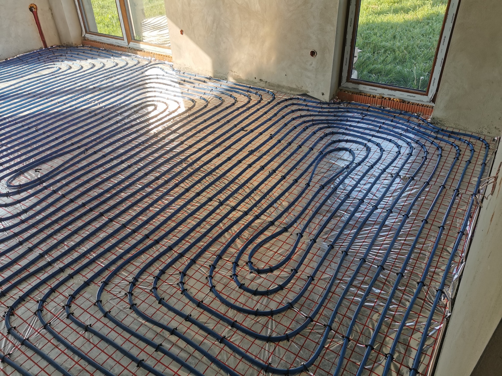 Niebieskie rurki ogrzewania podłogowego ułożone na folii z czerwoną siatką, widok na surową posadzkę i okno z widokiem na zieleń.