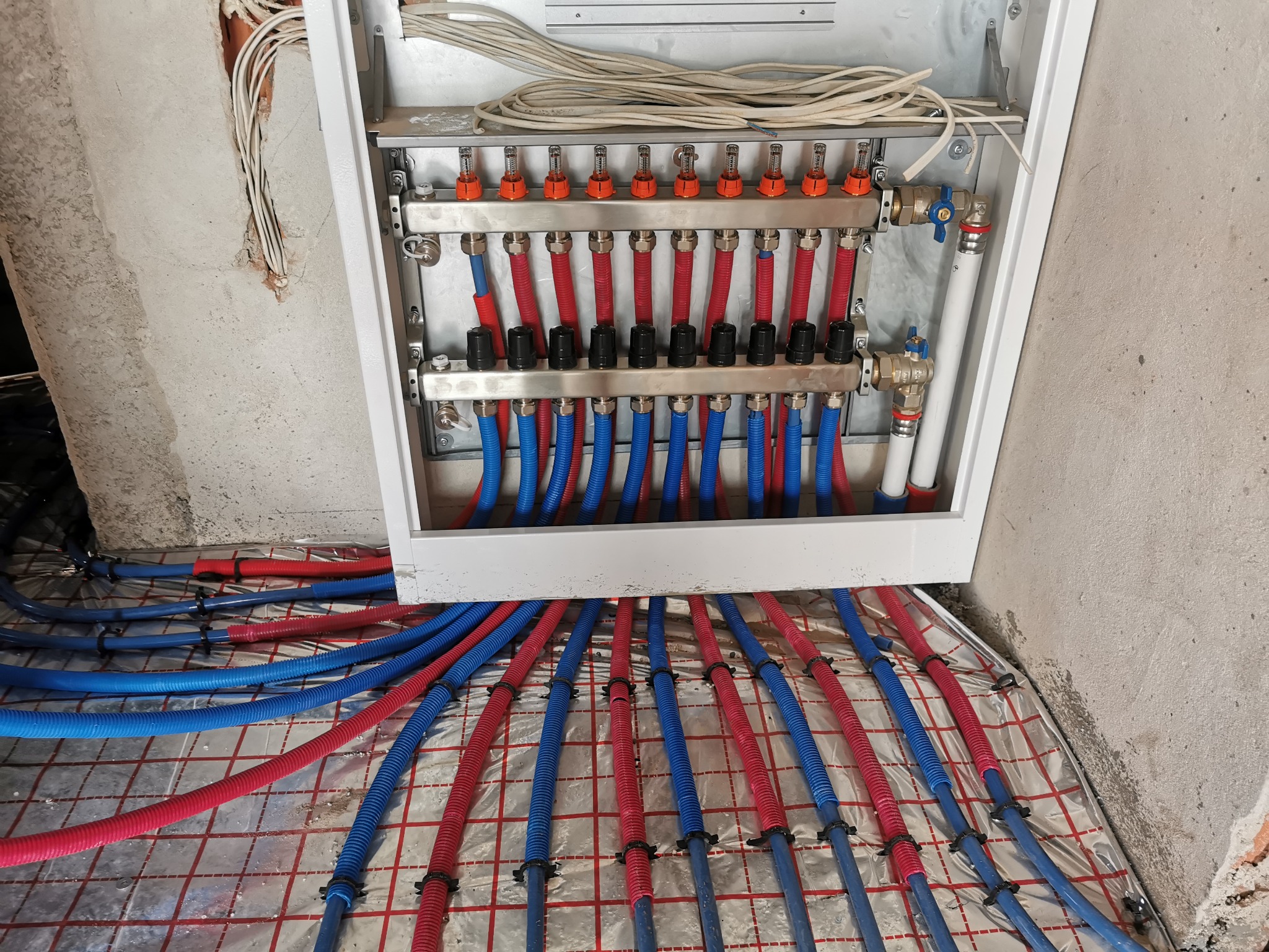 Rozdzielacz ogrzewania podłogowego z czerwonymi i niebieskimi rurami na folii izolacyjnej z siatką, widoczne zawory i odpowietrzniki, surowy beton w tle.