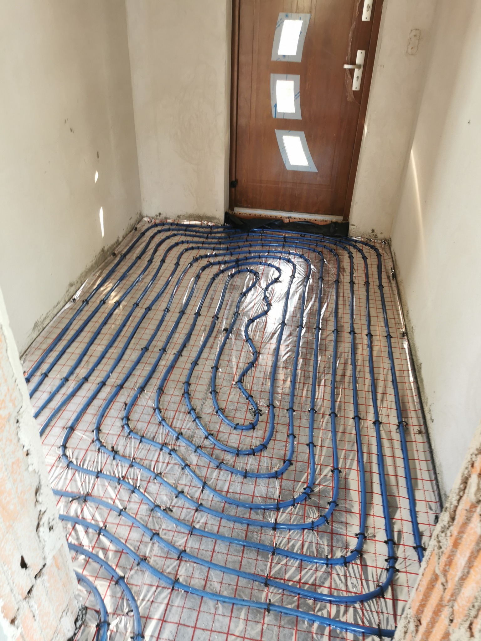 Niebieskie rury ogrzewania podłogowego, ułożone na folii z czerwoną siatką, zamocowane czarnymi spinkami, widoczne ściany w surowym tynku i brązowe drzwi wejściowe