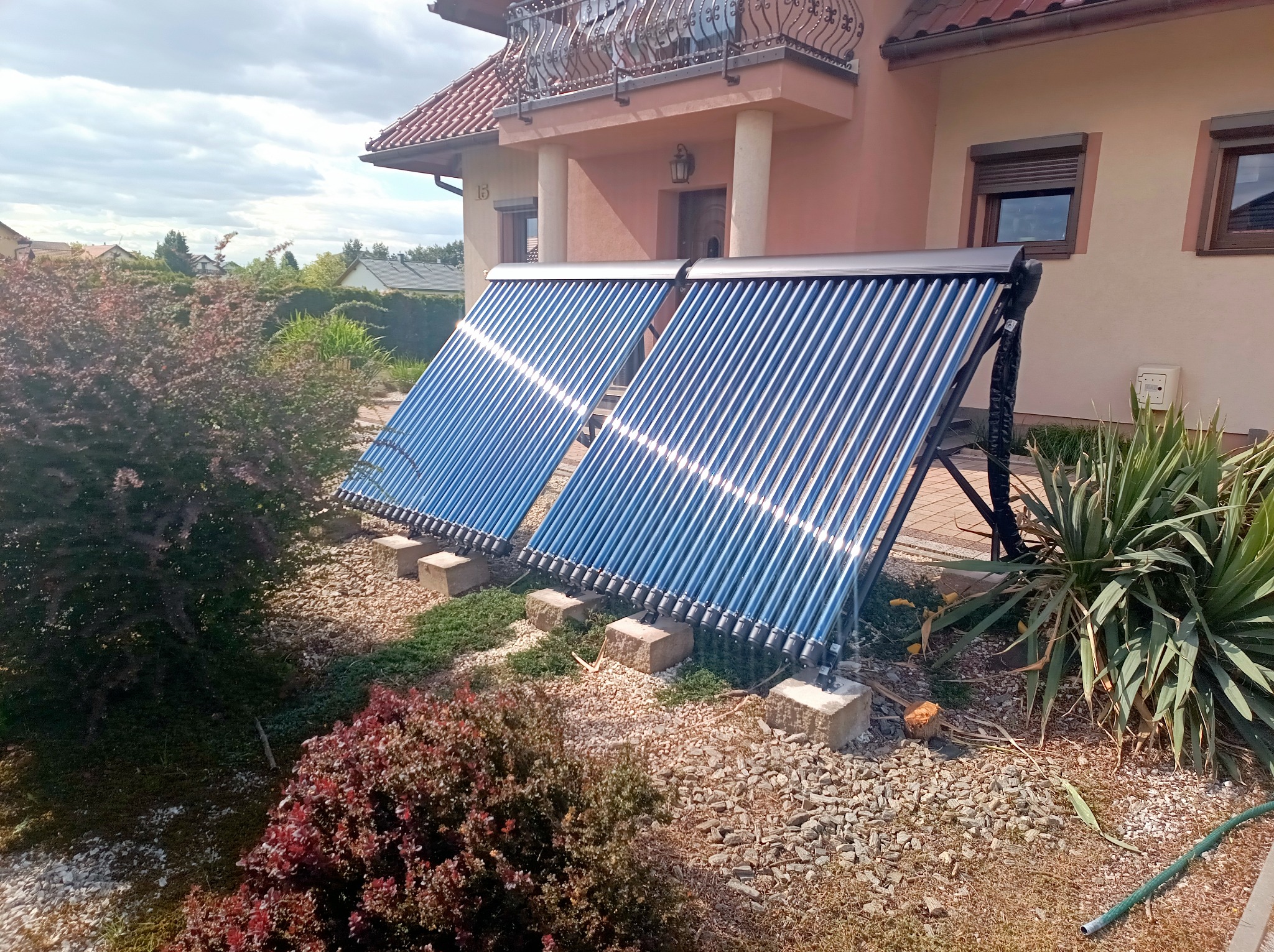 Dwa kolektory słoneczne rurowe zamontowane na zewnątrz domu, otoczone zielenią i kamieniami, z widocznym fragmentem elewacji budynku w tle.