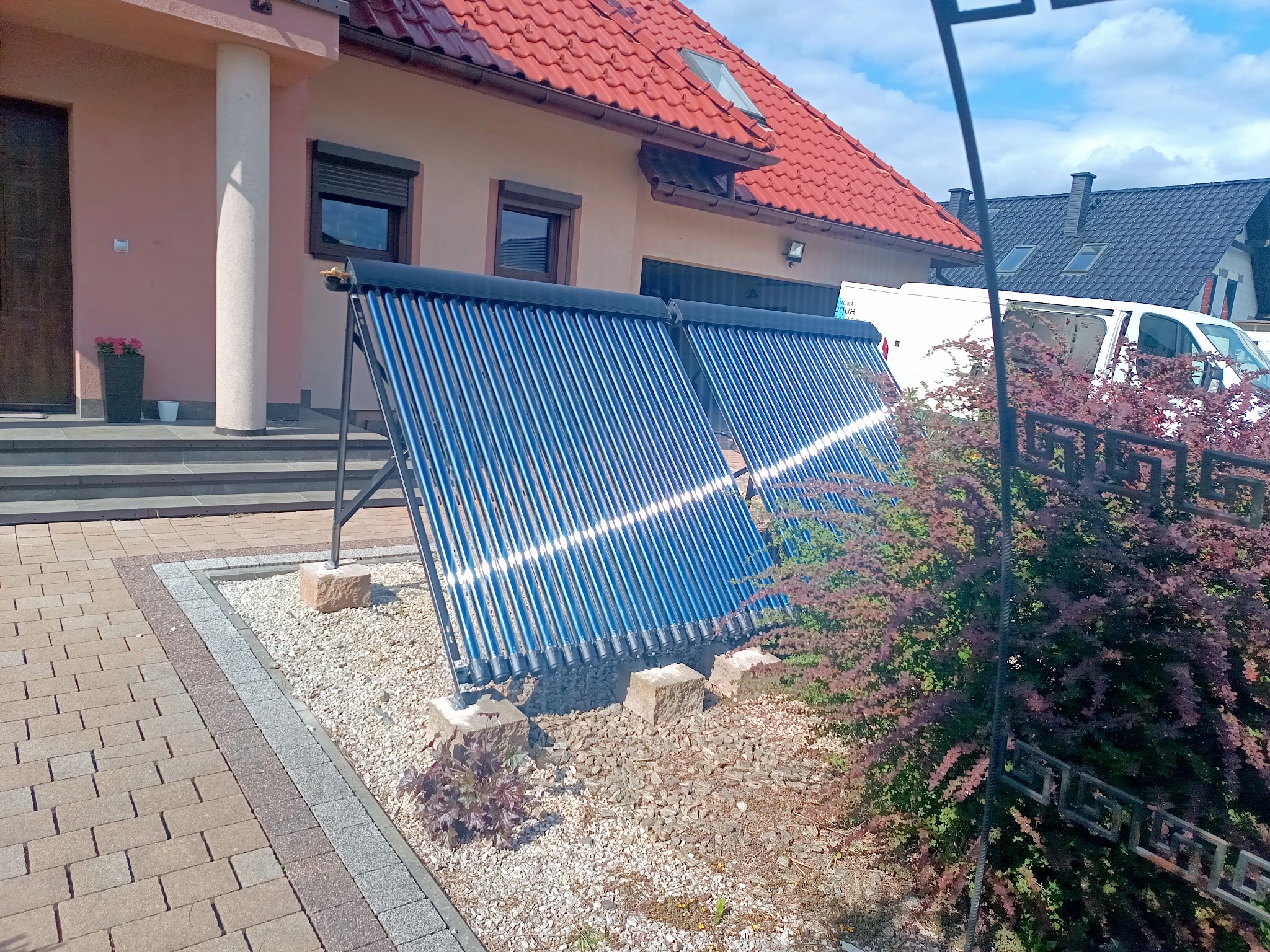 Zewnętrzny montaż kolektorów słonecznych na tle domu jednorodzinnego z czerwoną dachówką i ozdobnym brukiem. Widoczny biały bus. Słoneczny dzień.