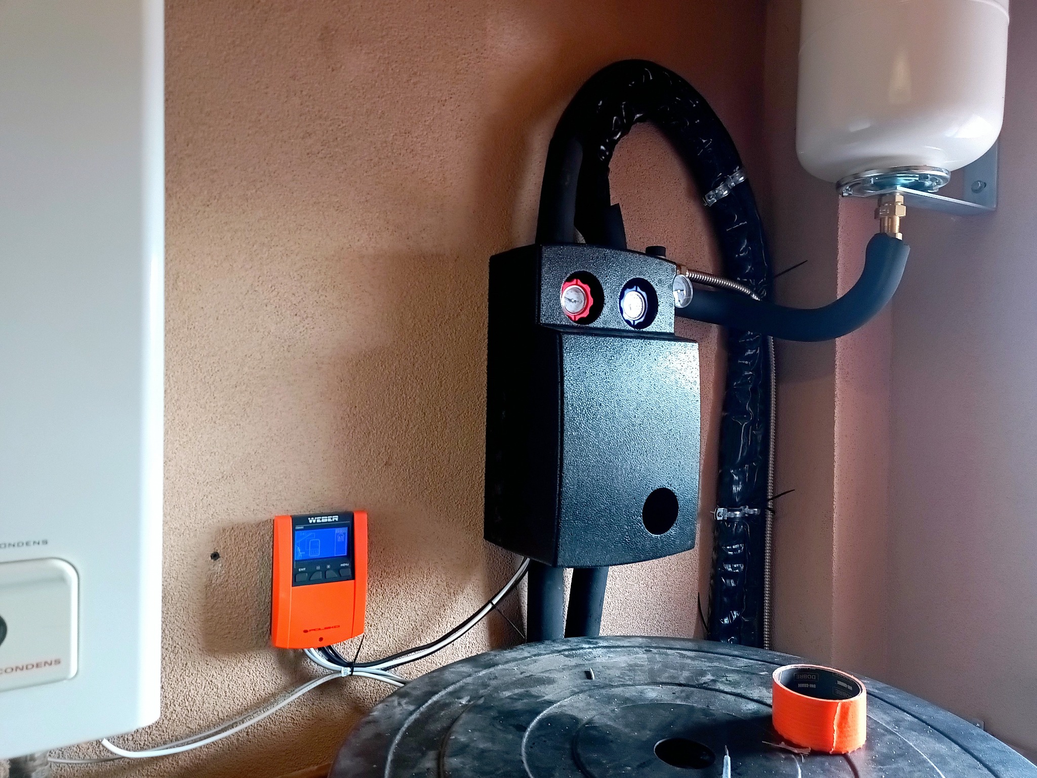 Zamontowany piec gazowy z regulatorem temperatury Weber i zbiornikiem. Czarna, izolowana rura doprowadzona do zbiornika. Widoczny pomarańczowy regulator na ścianie.