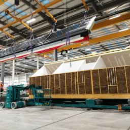 Roof-Dach - Prefabrykowany element dachu, podnoszony przez suwnicę w hali produkcyjnej, widoczny również element ściany z izolacją, pracownik w oddali.