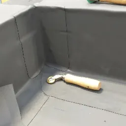 Szare pokrycie dachowe z detalami uszczelnienia narożnika, widoczny dociskowy wałek z drewnianą rączką oraz fragment arkusza materiału.