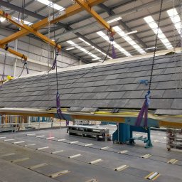 Roof-Dach - Prefabrykowany fragment dachu z szarymi dachówkami łupkowymi podnoszony przez suwnicę w hali produkcyjnej, widoczny pracownik w kasku i kamizelce odblaskowej.