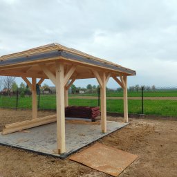 Roof-Dach - Budowa drewnianej altany z widocznym szkieletem dachu i stosem czerwonych dachówek w tle, na przygotowanym betonowym podłożu.