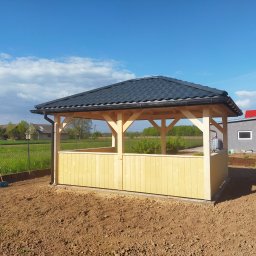 Roof-Dach - Nowo postawiona, jasna, drewniana altana z czarnym dachem z dachówki bitumicznej, częściowo obita deskami, na przygotowanym podłożu z widoczną szczotką i budynkami w tle.