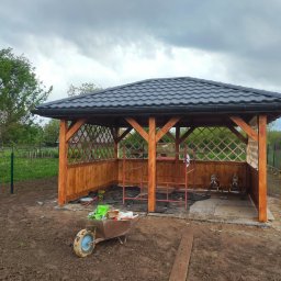 Roof-Dach - Altana z czarną dachówką w trakcie budowy, widoczne rusztowania wewnątrz konstrukcji, taczka z narzędziami i materiałami budowlanymi na pierwszym planie, ziemia wokół altany.