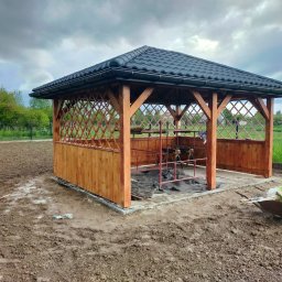 Roof-Dach - Drewniana altana z czarnym dachem z dachówki, w trakcie budowy, z widoczną konstrukcją, narzędziami i taczką na zewnątrz, na tle zieleni i pochmurnego nieba.