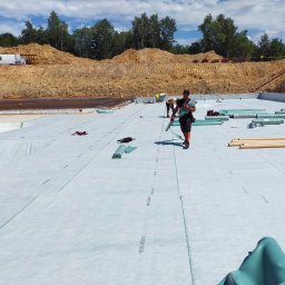 Roof-Dach - Przygotowanie podłoża pod taras: robotnicy rozkładają membranę izolacyjną na wyrównanym terenie budowy, w tle widoczny wykop i sprzęt budowlany.