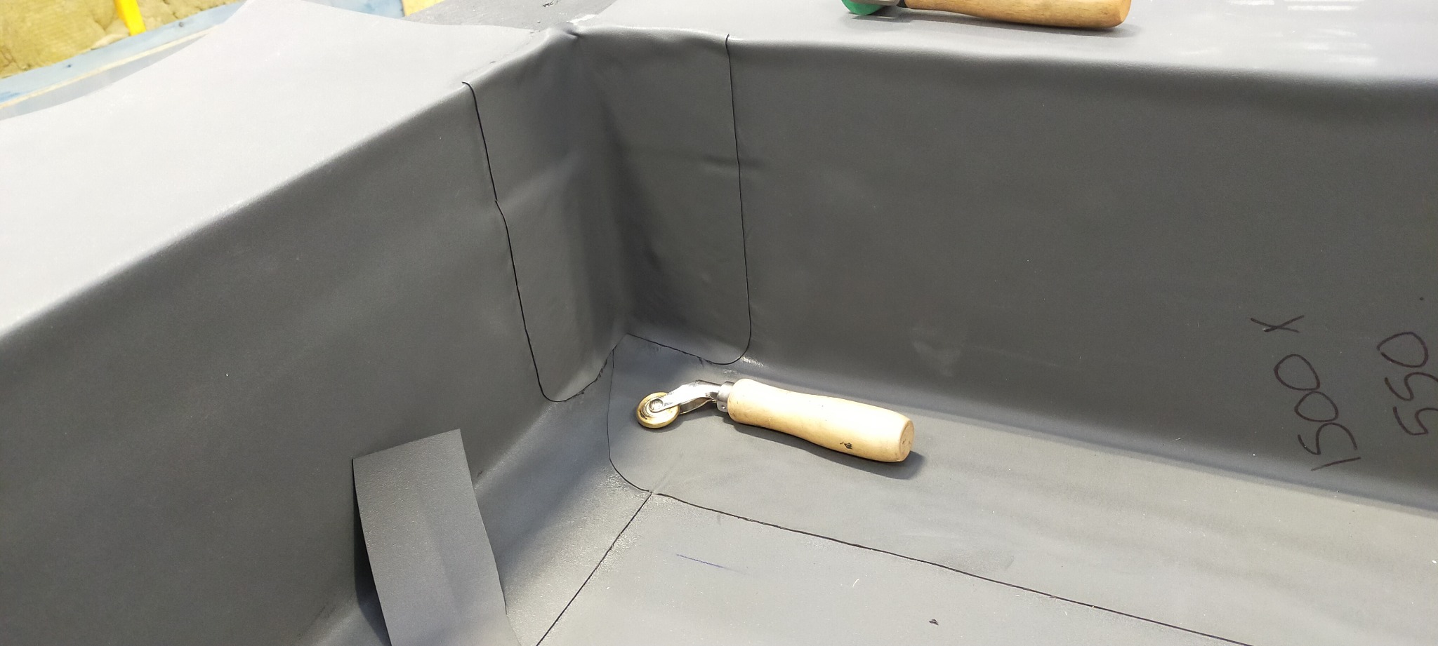 Szare pokrycie dachowe z detalami uszczelnienia narożnika, widoczny dociskowy wałek z drewnianą rączką oraz fragment arkusza materiału.