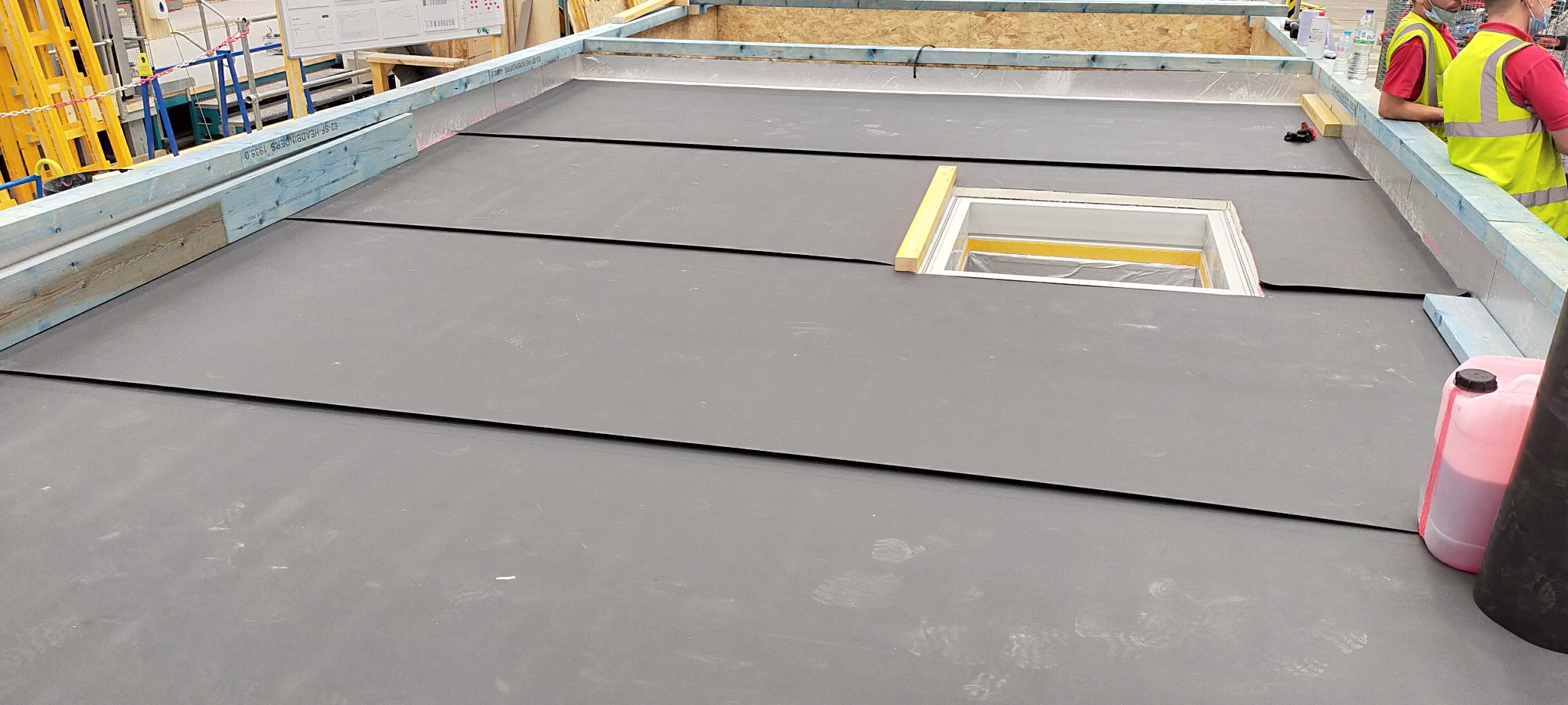 Montaż czarnej membrany dachowej z wycięciem na okno dachowe, widoczne drewniane elementy konstrukcyjne i fragmenty osób w odblaskowych kamizelkach.