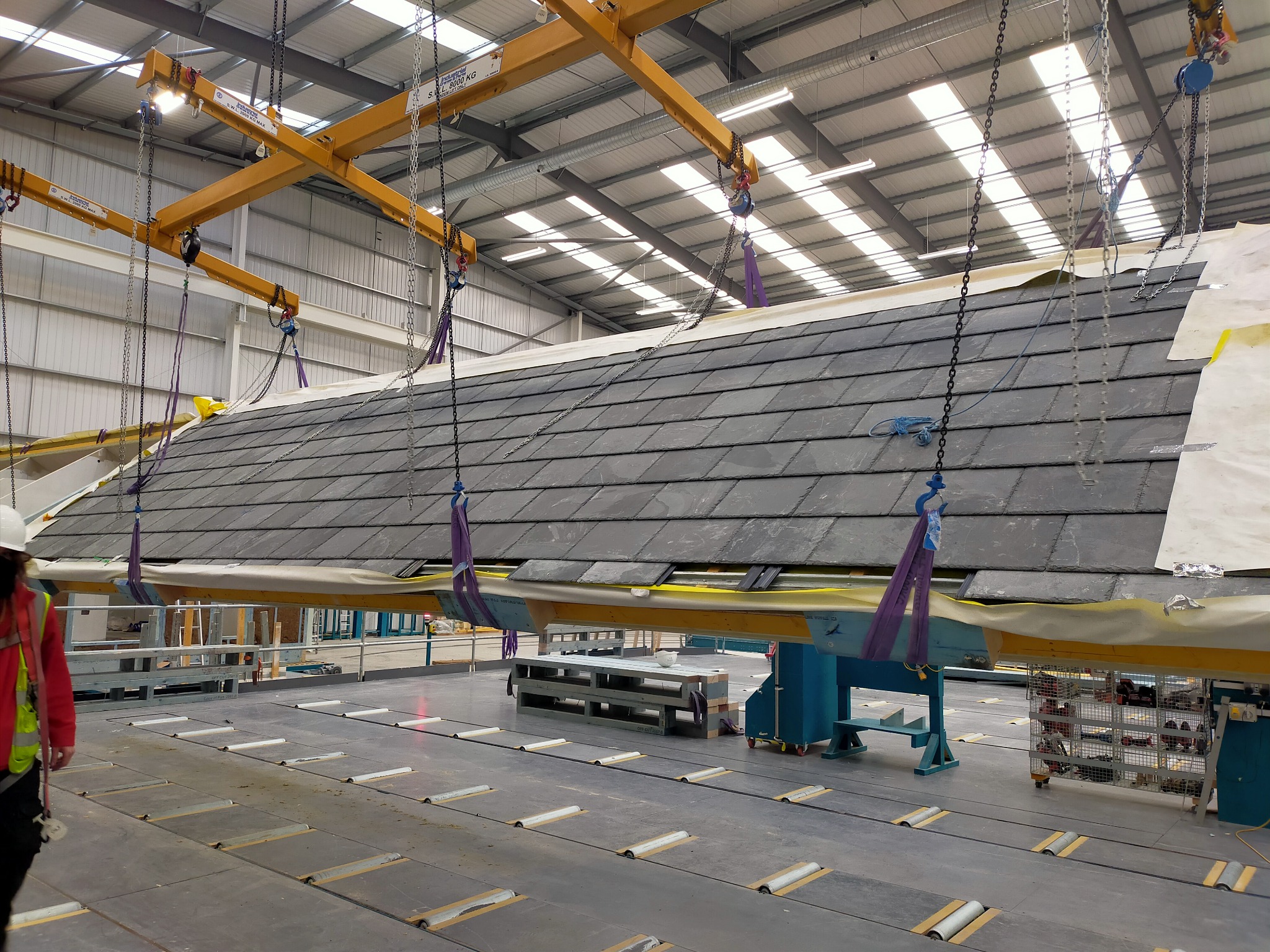 Prefabrykowany fragment dachu z szarymi dachówkami łupkowymi podnoszony przez suwnicę w hali produkcyjnej, widoczny pracownik w kasku i kamizelce odblaskowej.