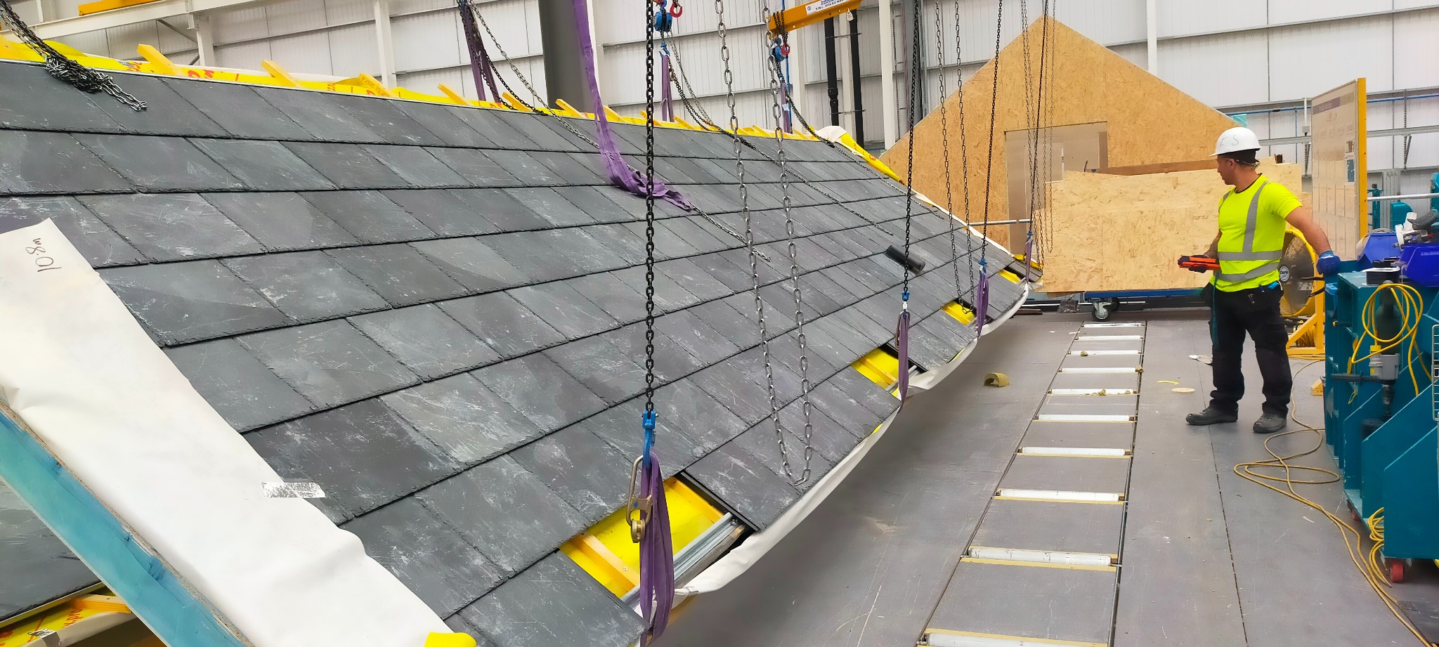 Fragment dachu z szarej dachówki łupkowej, podnoszony za pomocą łańcuchów w hali produkcyjnej; widoczny pracownik w kasku i kamizelce odblaskowej.
