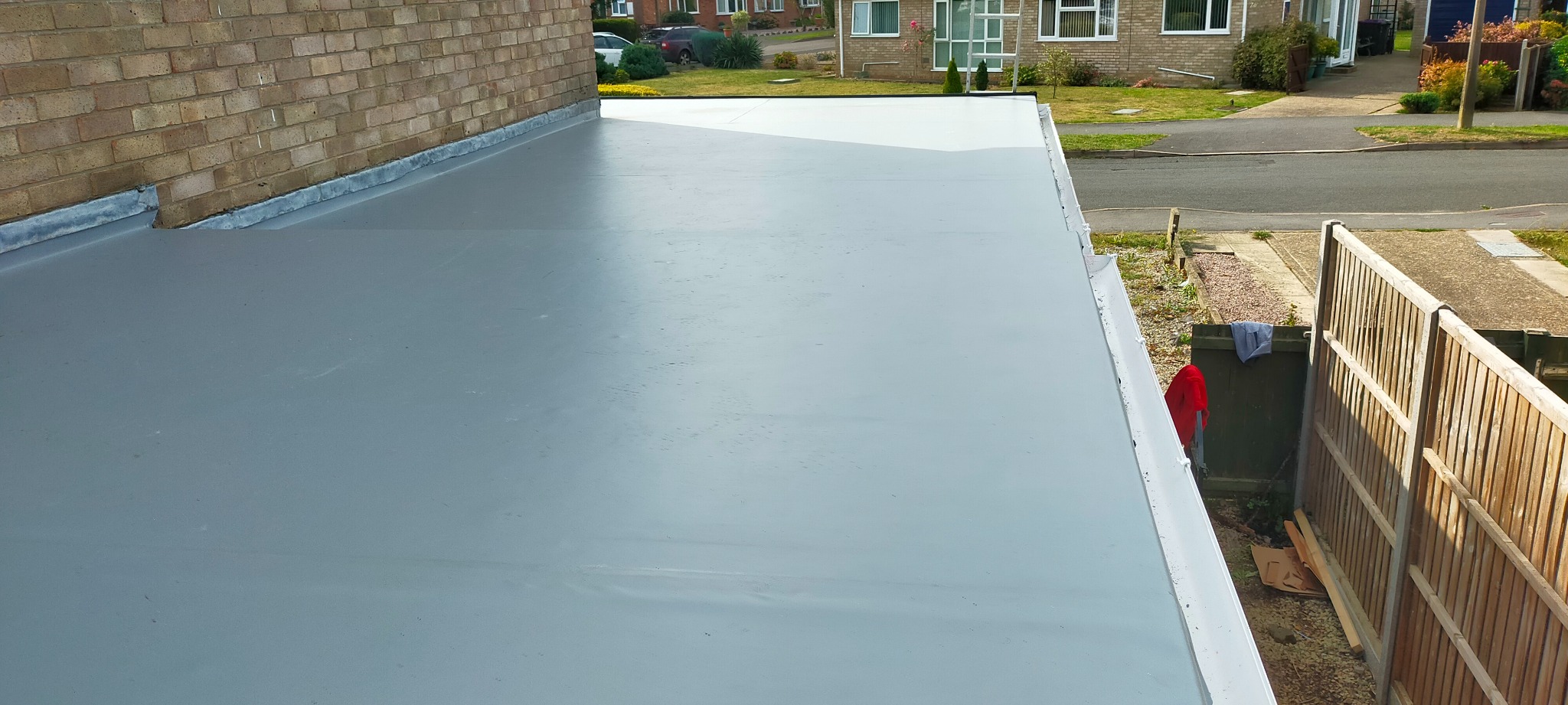 Szare, płaskie pokrycie dachu z widocznymi detalami wykończenia przy ścianie budynku i rynną, otoczone zielenią i zabudową mieszkalną.