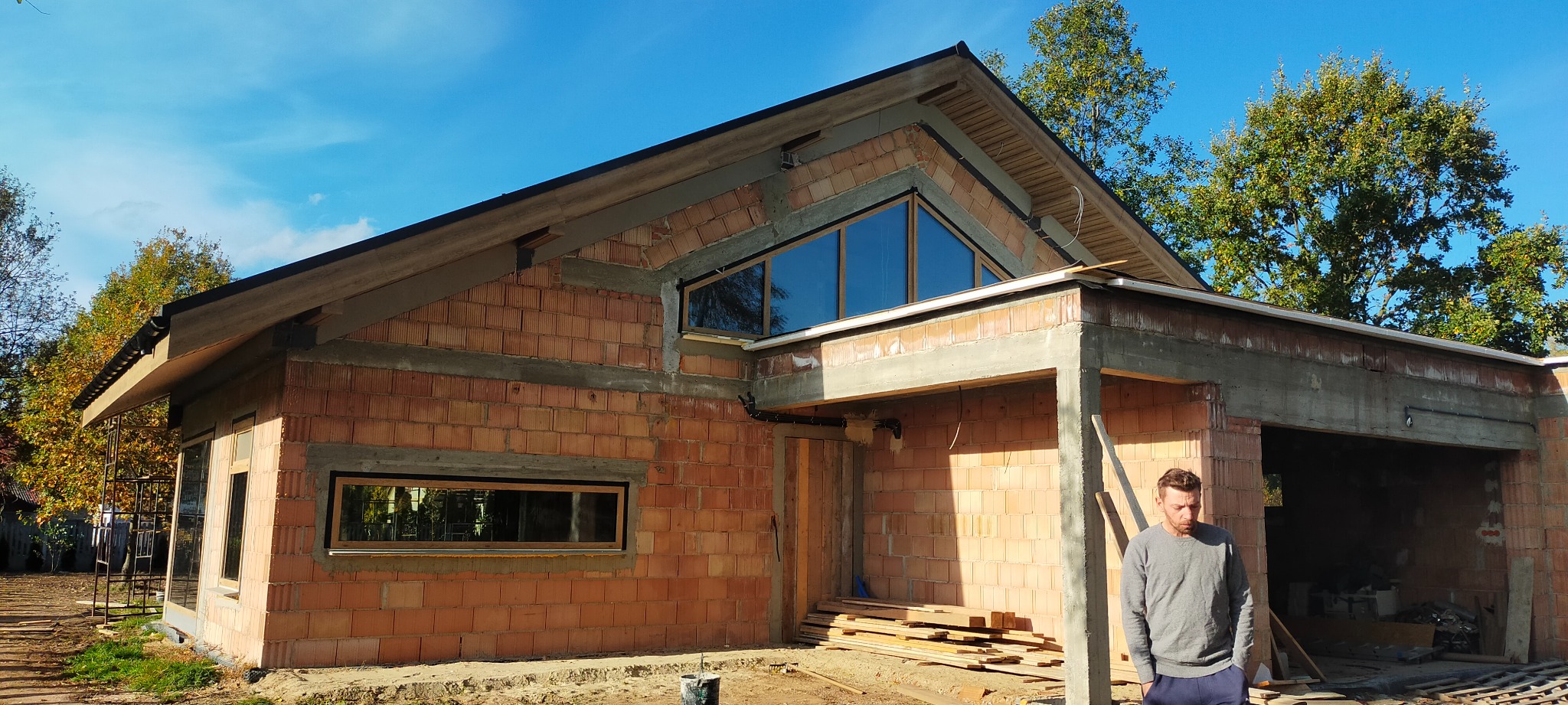 Budynek w trakcie budowy z widoczną konstrukcją dachu, ścianami z czerwonej cegły, oknami i mężczyzną w szarej bluzie stojącym przed budynkiem.