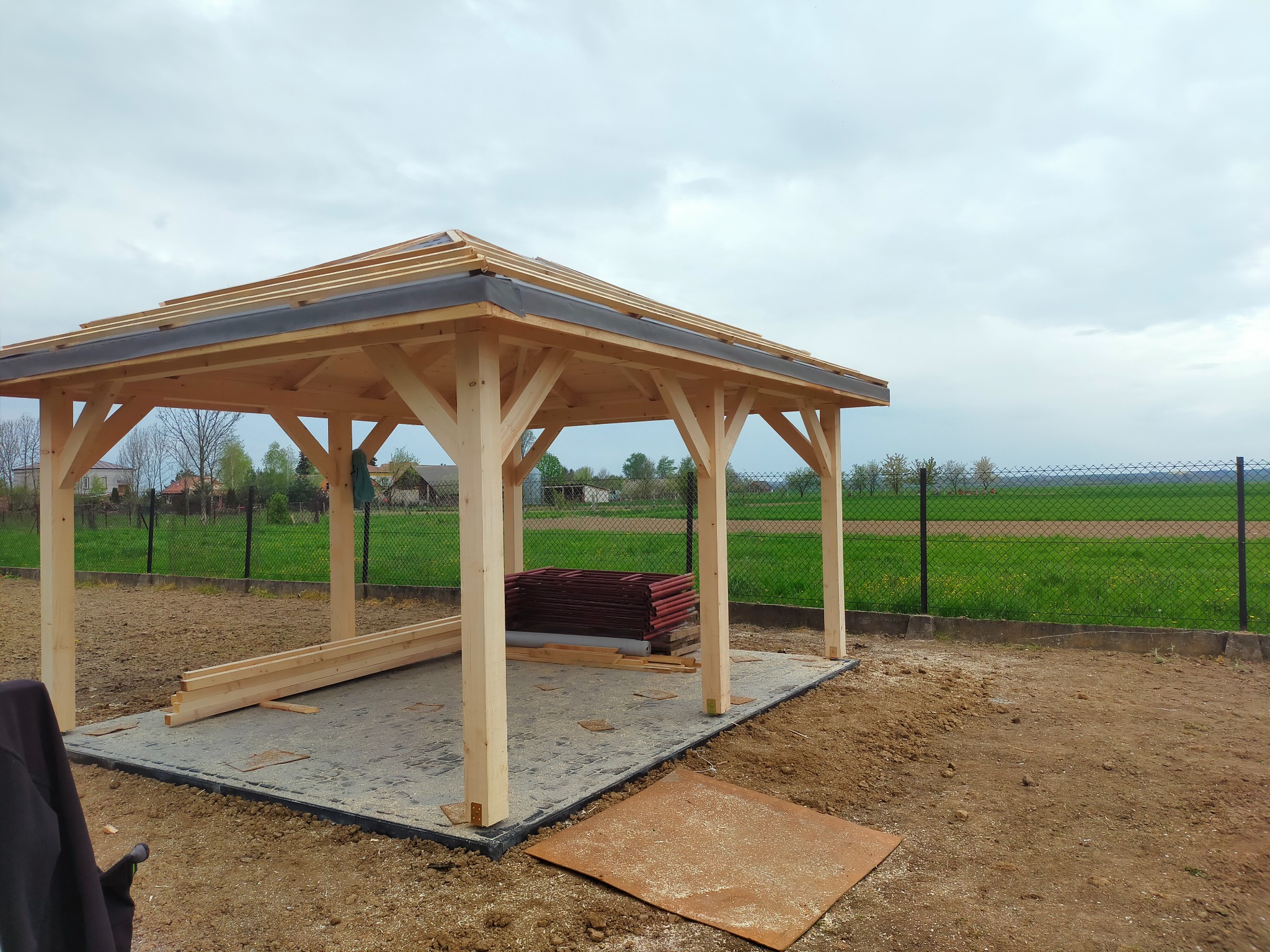 Budowa drewnianej altany z widocznym szkieletem dachu i stosem czerwonych dachówek w tle, na przygotowanym betonowym podłożu.