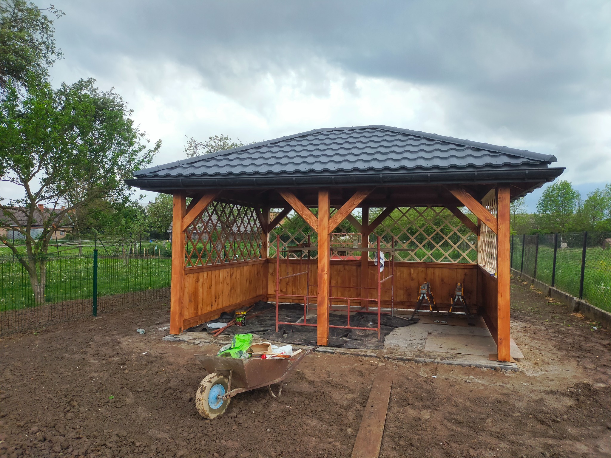 Altana z czarną dachówką w trakcie budowy, widoczne rusztowania wewnątrz konstrukcji, taczka z narzędziami i materiałami budowlanymi na pierwszym planie, ziemia wokół altany.