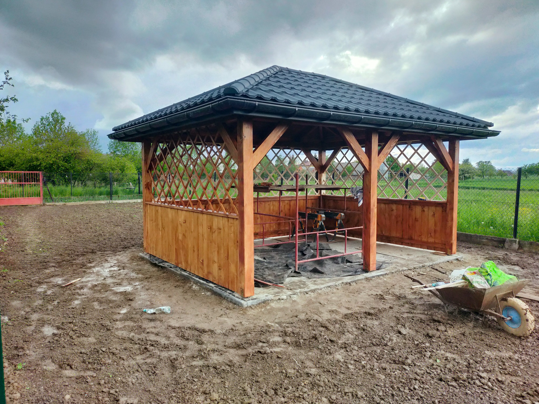Drewniana altana z czarnym dachem z dachówki, w trakcie budowy, z widoczną konstrukcją, narzędziami i taczką na zewnątrz, na tle zieleni i pochmurnego nieba.