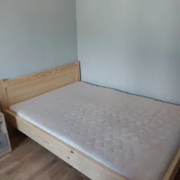 Surowe drewniane łóżko z materacem w pokoju z szarą ścianą i drewnianą podłogą, widok z boku.