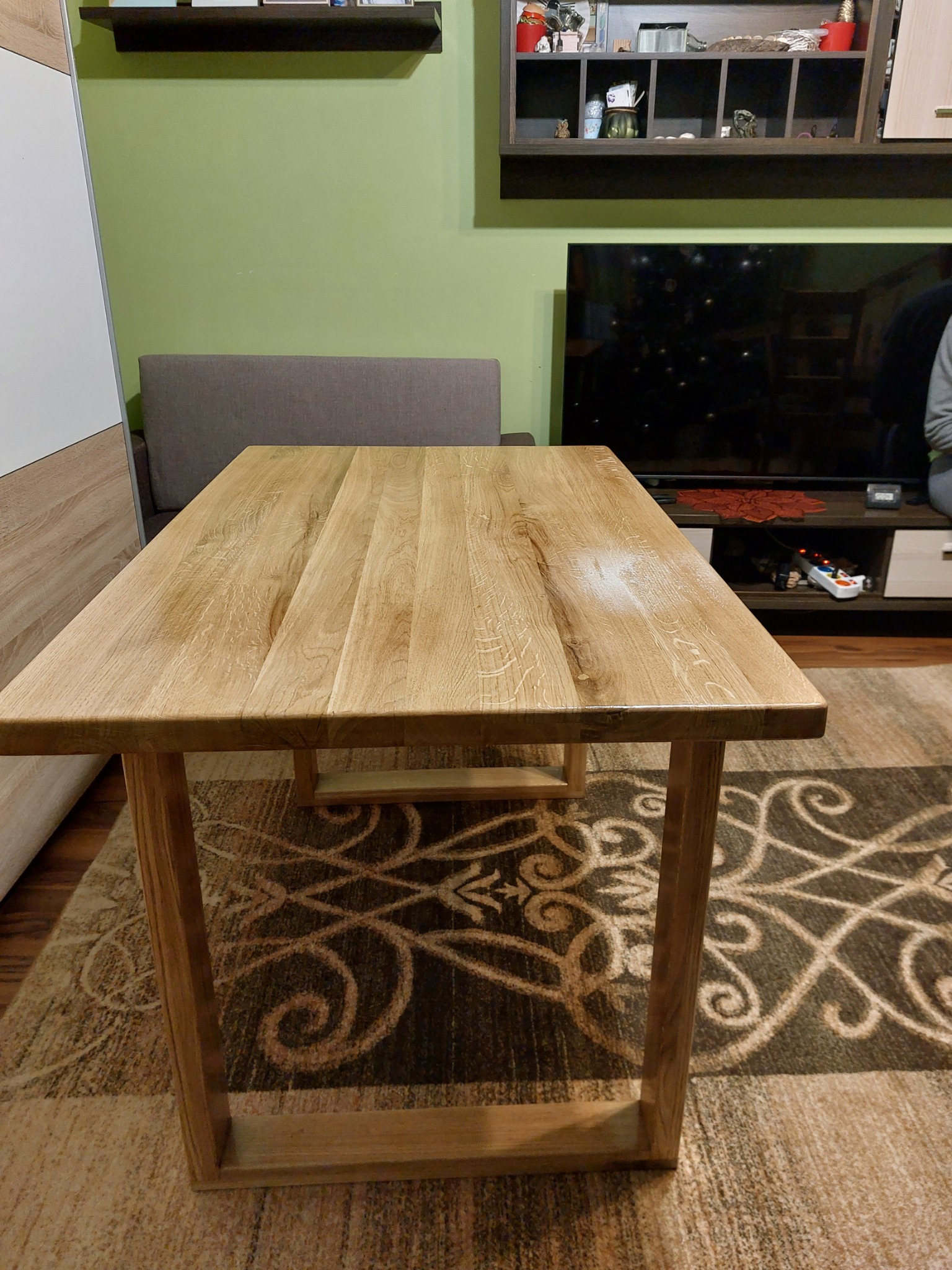 Stół z jasnego drewna o prostokątnym blacie i minimalistycznej konstrukcji, ustawiony na wzorzystym dywanie w salonie z zieloną ścianą i meblami w tle.