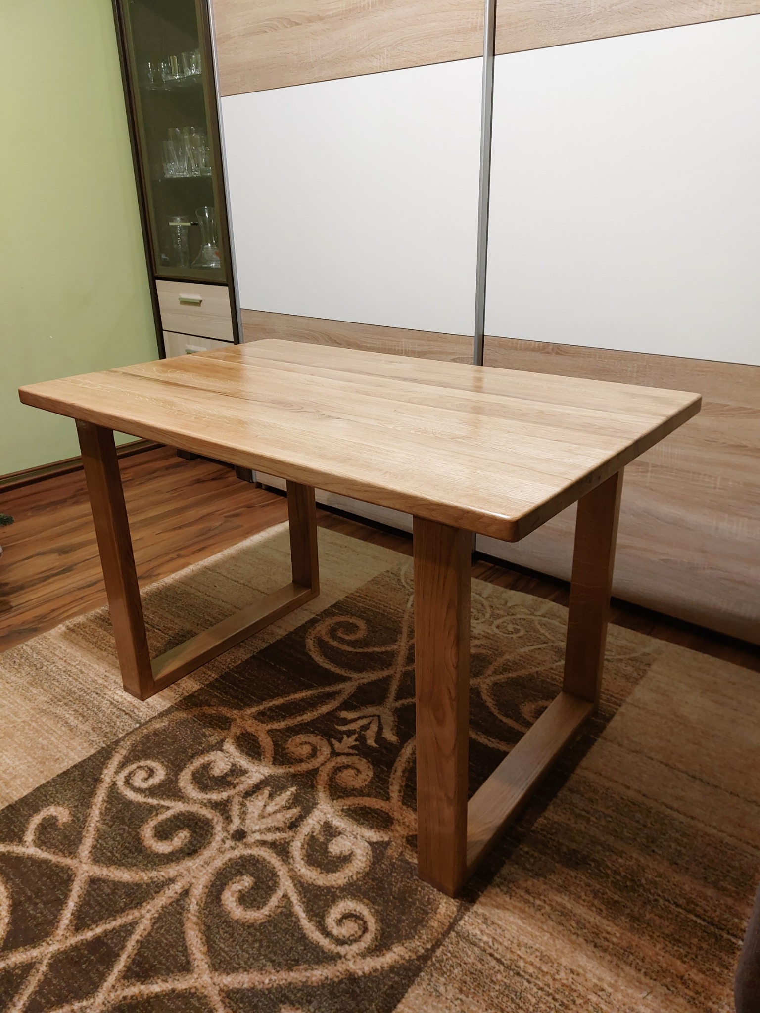 Jasny, drewniany stół o prostokątnym blacie i nogach w kształcie kwadratowych ram, stojący na wzorzystym dywanie w salonie z szafą w tle.