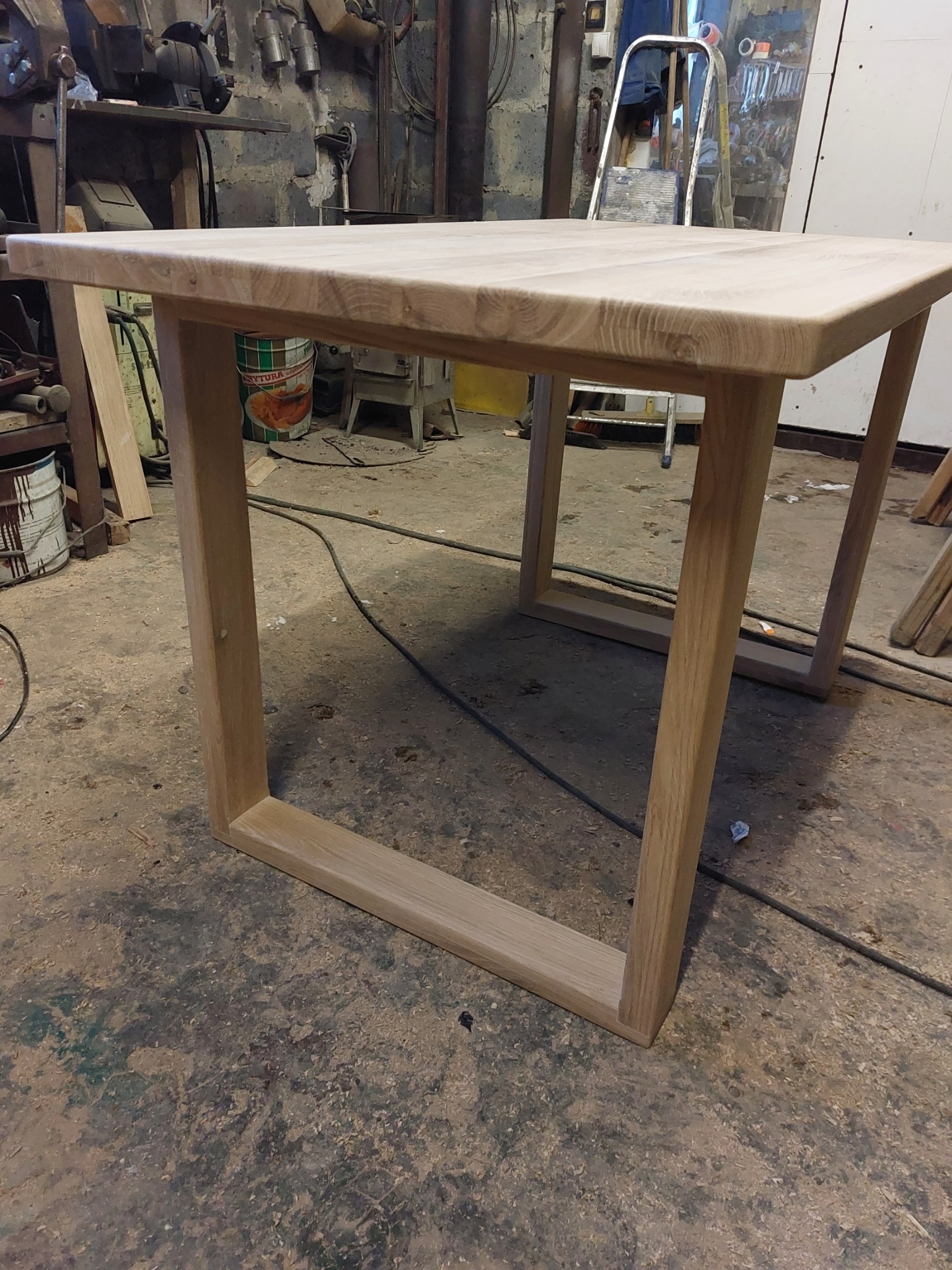 Surowy, drewniany stół z wyraźnym usłojeniem, w warsztacie z widocznym bałaganem na podłodze i narzędziami w tle.