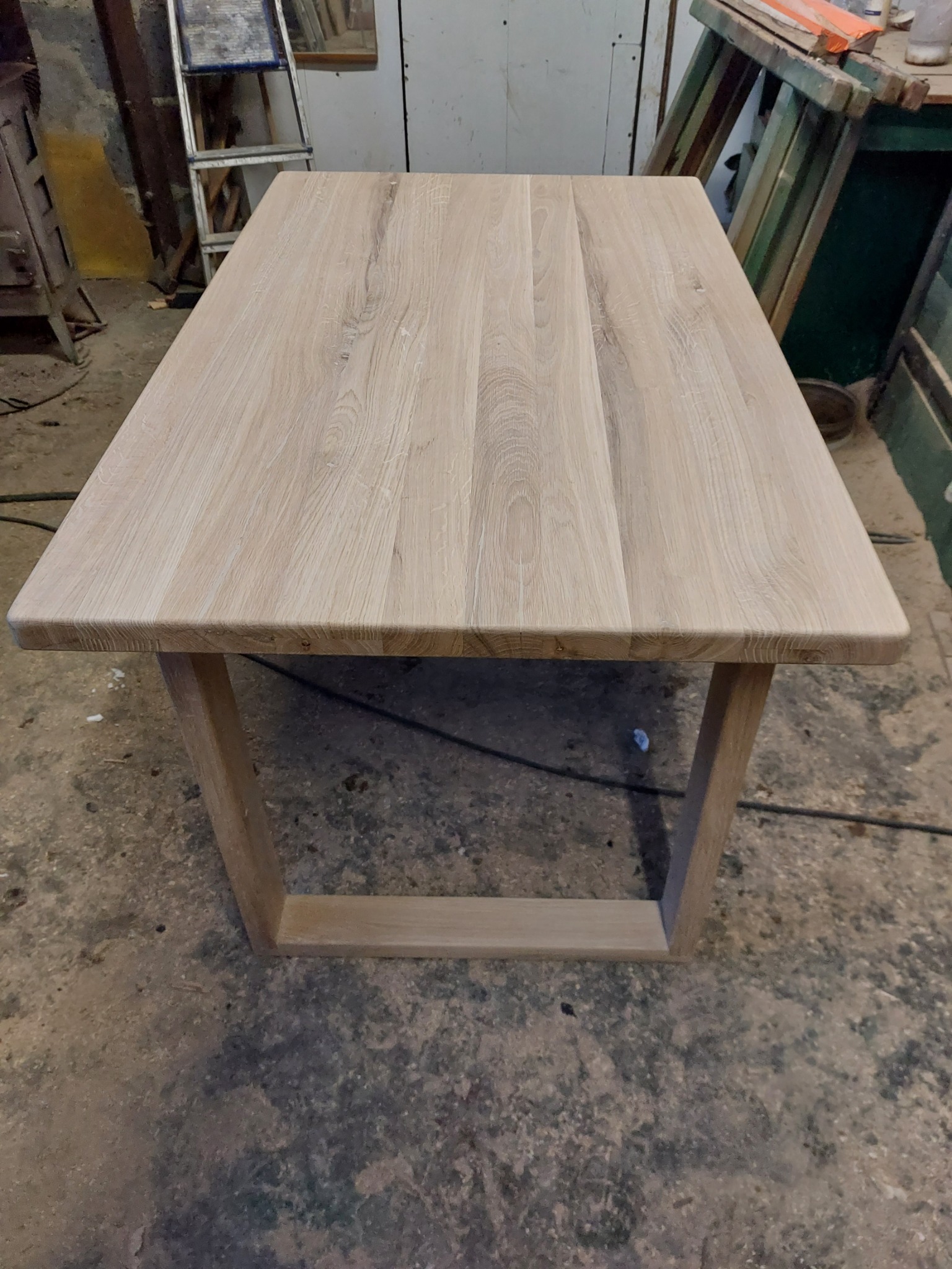 Surowy, drewniany stół z jasnego drewna w warsztacie, widoczne niedoskonałości podłogi i drabina w tle.