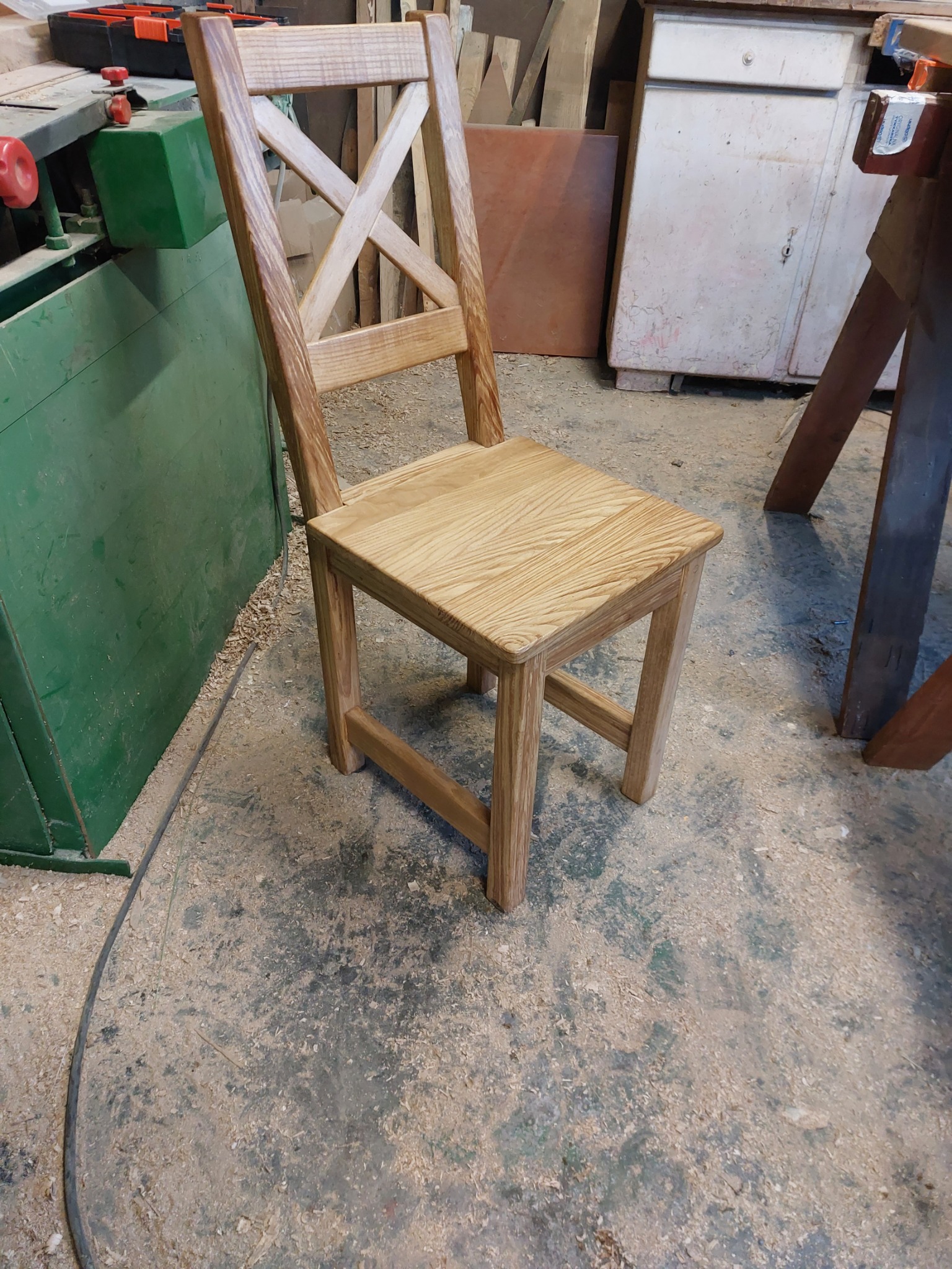 Surowe drewniane krzesło z krzyżowym oparciem stoi w warsztacie, otoczone trocinami i narzędziami.