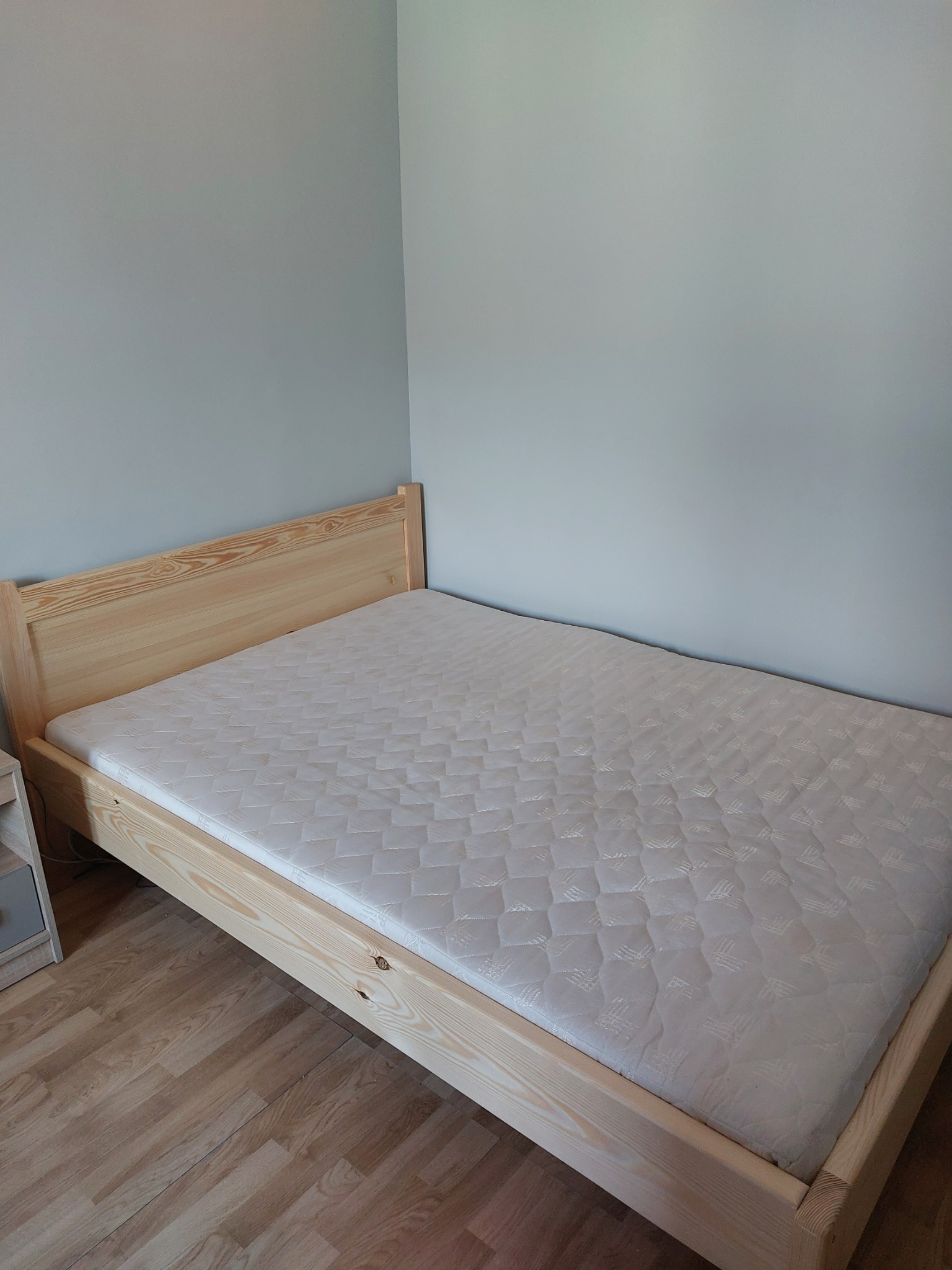 Surowe drewniane łóżko z materacem w pokoju z szarą ścianą i drewnianą podłogą, widok z boku.