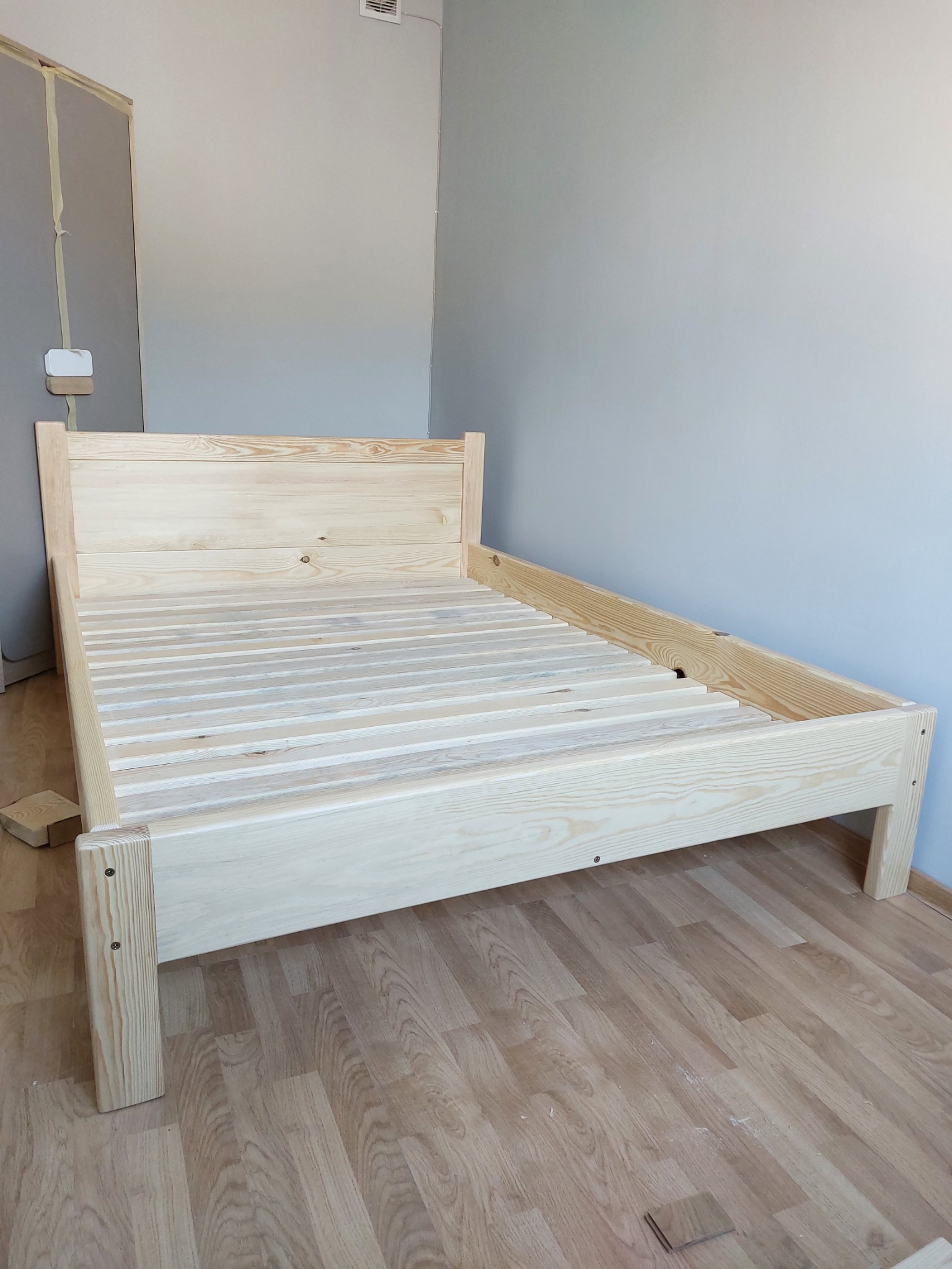 Surowa, drewniana rama łóżka z listewkami na podłodze z paneli, widoczne ślady montażu i fragmenty drewna, szara ściana w tle.