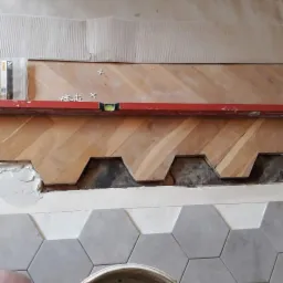 Fragment podłogi w trakcie układania: heksagonalne szare płytki ceramiczne przechodzą w ułożony parkiet w jodełkę, z widocznym poziomem, krzyżykami dystansowymi i przygotowanym podłożem.