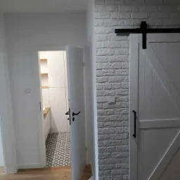 Korytarz z widokiem na łazienkę, biała cegła na ścianie i przesuwne drzwi stodołowe z czarnym okuciem.