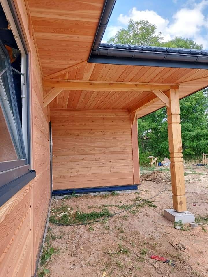 Fragment elewacji drewnianego domu w stanie surowym, widoczne okno z czarną ramą, drewniana kolumna podtrzymująca okap i czarna rynna, ziemiste podłoże.