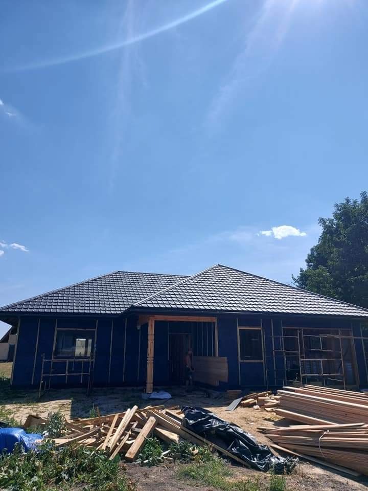 Niedokończony dom z czarnym dachem i niebieską elewacją, widoczne drewniane elementy konstrukcyjne, przygotowane deski na pierwszym planie, jasne niebo w tle.