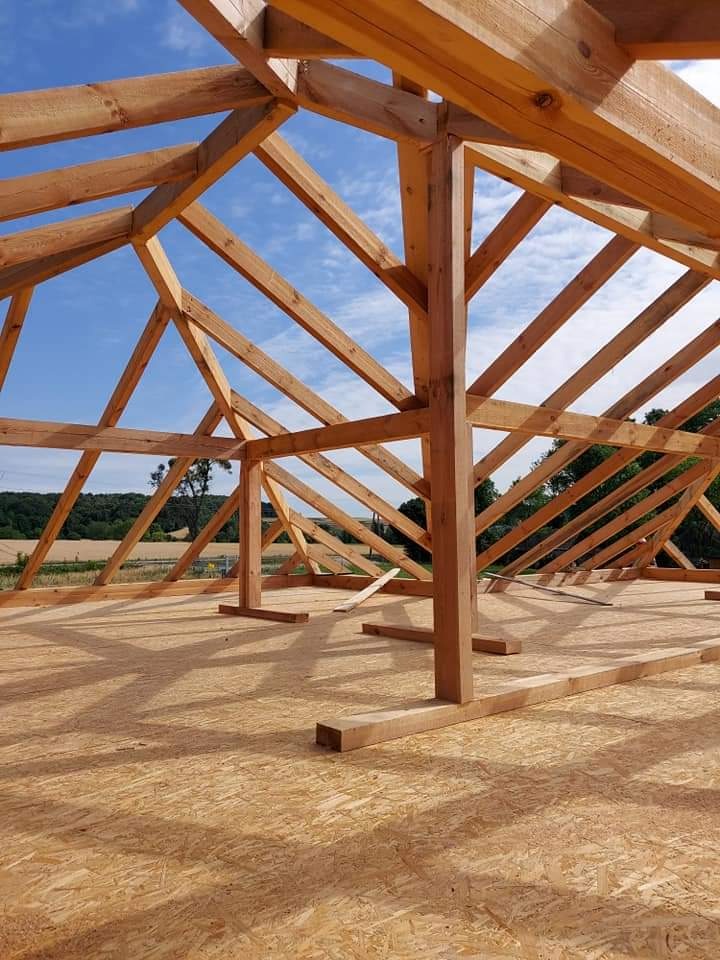 Konstrukcja drewniana domu w trakcie budowy, widoczna więźba dachowa z surowego drewna na tle błękitnego nieba i zieleni