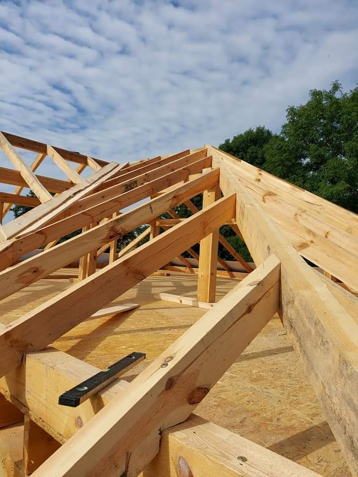 Drewniana konstrukcja dachu w trakcie budowy, widoczne elementy więźby dachowej i poziomica na belce, na tle błękitnego nieba z chmurami.