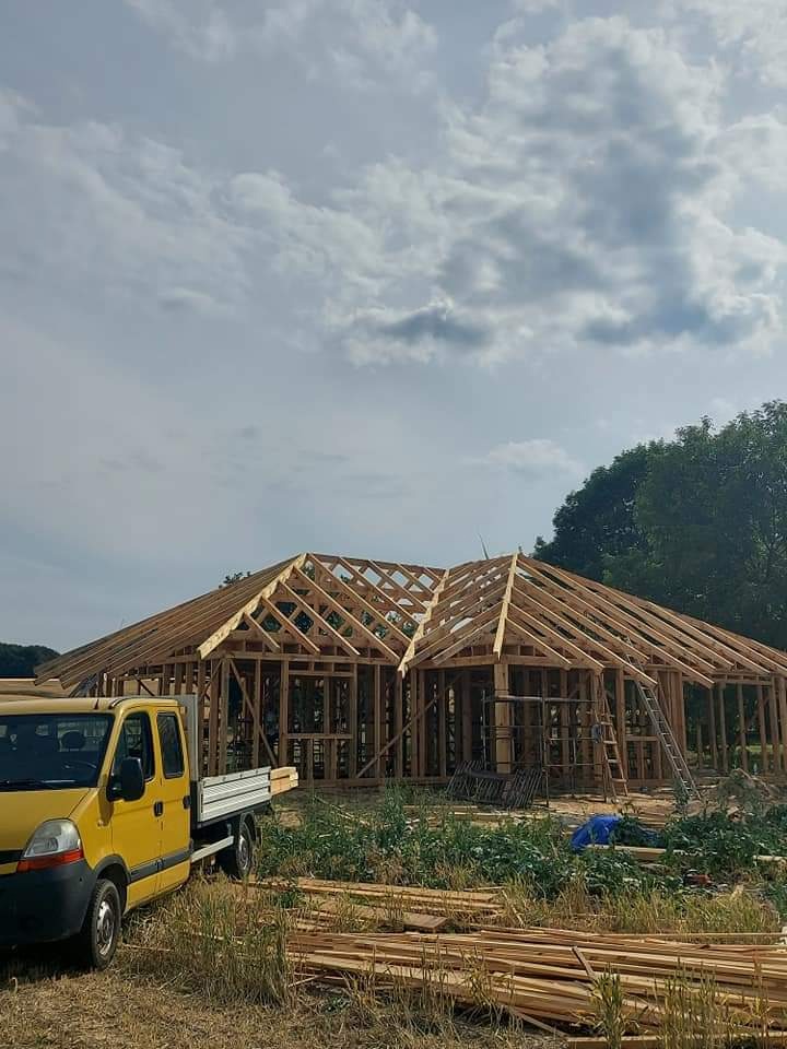 Budowa domu w konstrukcji szkieletowej z widoczną więźbą dachową, żółty samochód dostawczy w tle, plac budowy z drewnem i rusztowaniami.