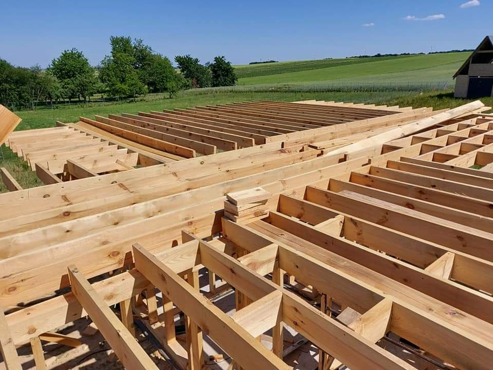 Wiązar dachowy z jasnego drewna w trakcie montażu, widok z góry na tle zielonych pól i błękitnego nieba.